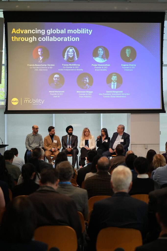 A wide shot of a panel discussion titled "Advancing global mobility through collaboration" taking place at the MaRS Toyota Mobility Forum. Seven panelists are seated on stage, facing a large, engaged audience. A screen above them displays their names and titles, including Tracey McGillivray of Axtion Independence Mobility, Pooja Viswanathan of Braze Mobility, and Manmeet Maggu of Trexo Robotics. The moderator, David Hengeveld, is seated on the far right.