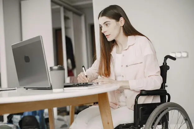 A young woman in a wheelchair working at a light-colored table, looking at a laptop and writing notes in a planner.