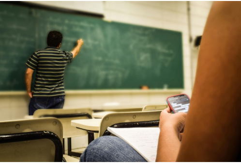 Student in the foreground looking at a cell phone during a class, while a teacher writes on a green chalkboard in the background.