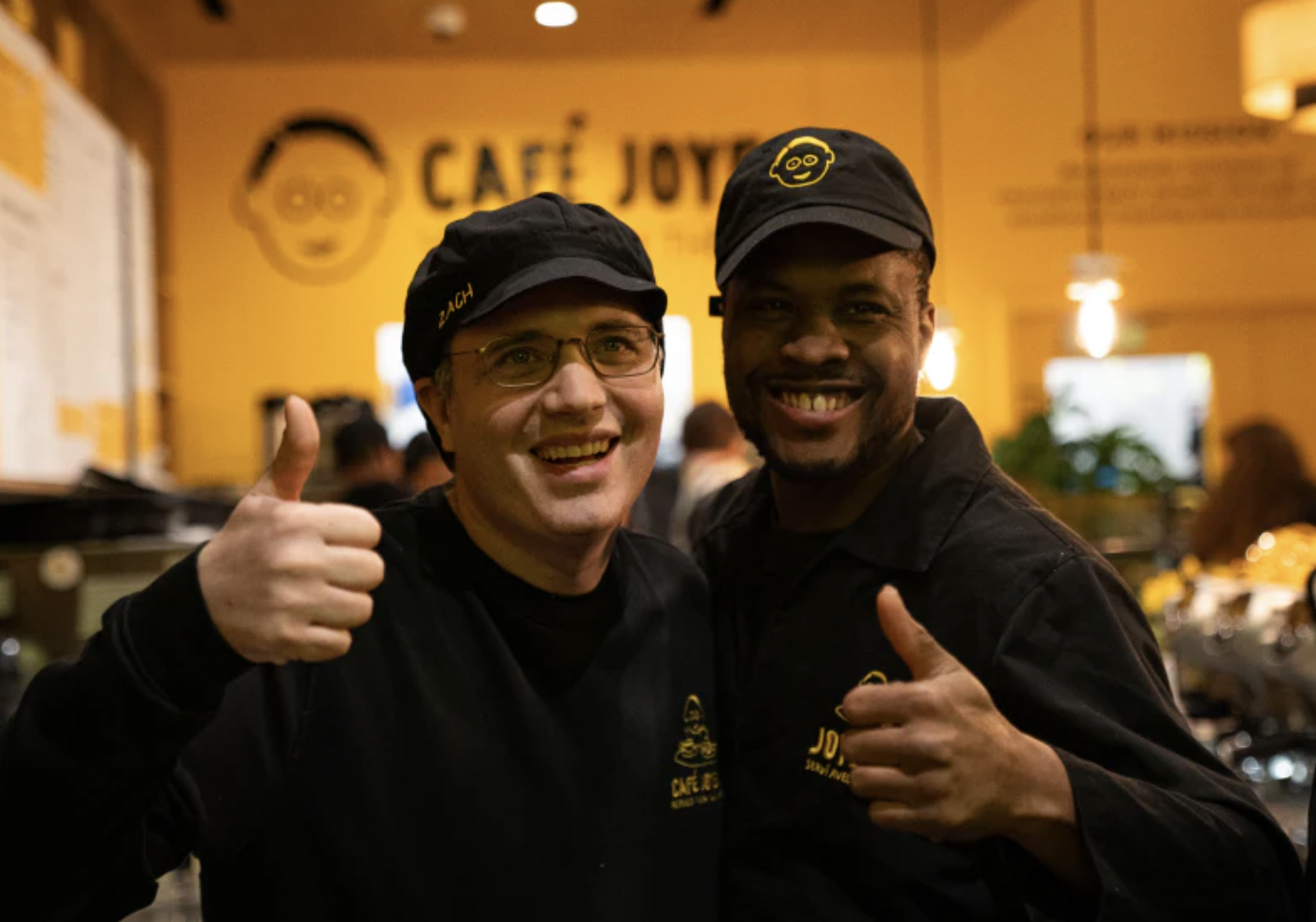 Two men, both wearing black shirts and black caps, stand together smiling brightly and giving thumbs up gestures in what appears to be a cafe. The man on the left wears glasses. The background is a bright yellow wall with the words "CAFÉ JOYEUX" and a stylized face logo written on it in black.