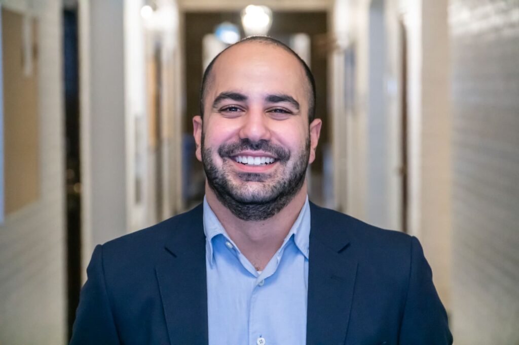 A professional headshot of Emile Maamary. He is a bald man with a dark beard, wearing a navy blue suit jacket over a light blue collared shirt. He is smiling widely and looking directly at the camera. He is standing in a brightly lit hallway with a blurred background of architectural details.
