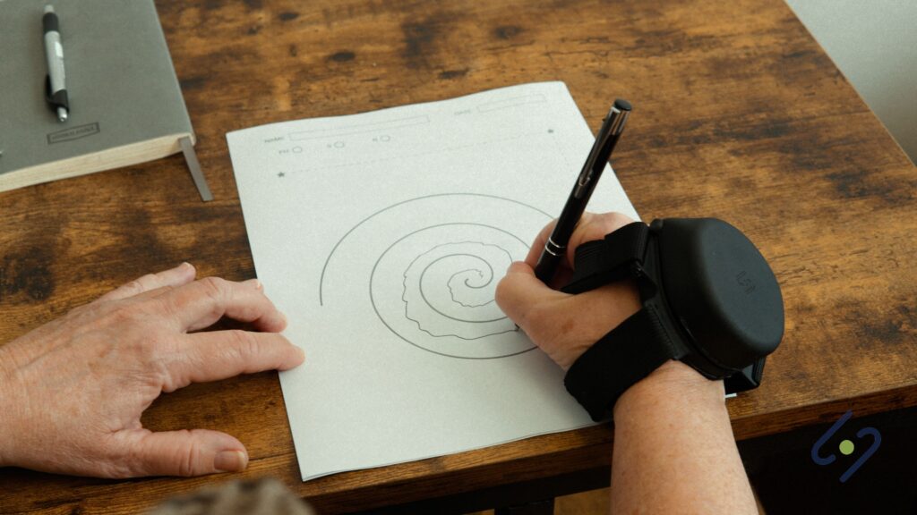 A close-up, overhead shot of a person's hands performing a spiral drawing test, commonly used to assess tremor severity. The person's writing hand is stabilized by a black wrist-worn device (likely a Steadi-One or similar anti-tremor orthosis) as they use a pen to trace a spiral pattern on a piece of paper. The other hand rests on the wooden table. A notebook is visible in the background.