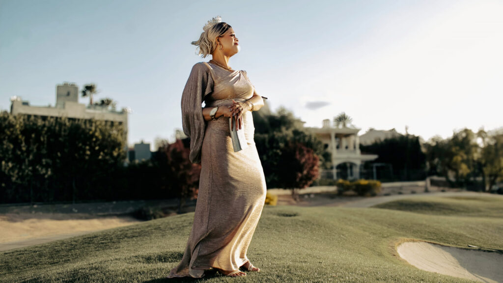 A woman with blonde hair, wearing a crown and a flowing, metallic gold dress, stands outdoors on a manicured, sun-drenched golf course or park, looking into the distance.