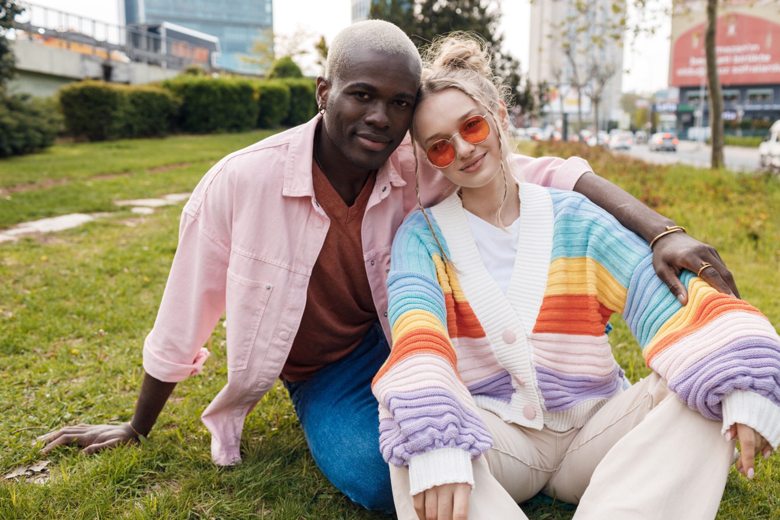 A smiling couple sitting together on a grassy patch in an urban park. The Black man on the left has short, platinum blonde hair and is wearing a pink denim jacket over a terracotta shirt and blue jeans. He has his arm around the white woman on the right, who is wearing a colorful striped cardigan (pink, yellow, orange, blue, and purple) over a white top and tan pants. She is wearing orange-tinted sunglasses and leaning her head against him. Cars and city buildings are visible in the soft-focus background.