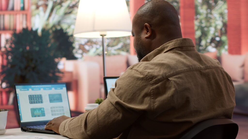 A rear, over-the-shoulder view of a Black man with a shaved head and beard, wearing a tan or light brown button-down shirt, working intently on a laptop at a desk in a well-lit home office or living space. The laptop screen displays a project dashboard with charts and data widgets. A large lamp with a white shade is visible behind him, and the background is softly focused, showing plush furniture and large plants, suggesting a comfortable, indoor-outdoor feel.