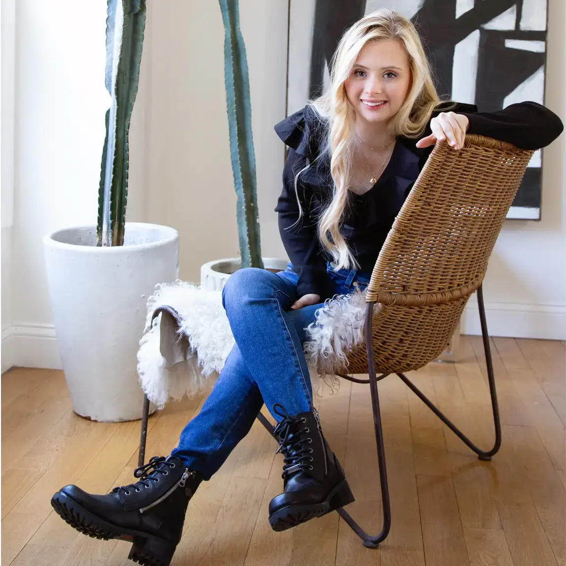 A blonde woman with Down syndrome sitting on a woven chair with a white sheepskin throw, wearing a black ruffled top, jeans, and black combat boots, smiling at the camera with large cacti and modern art in the background.