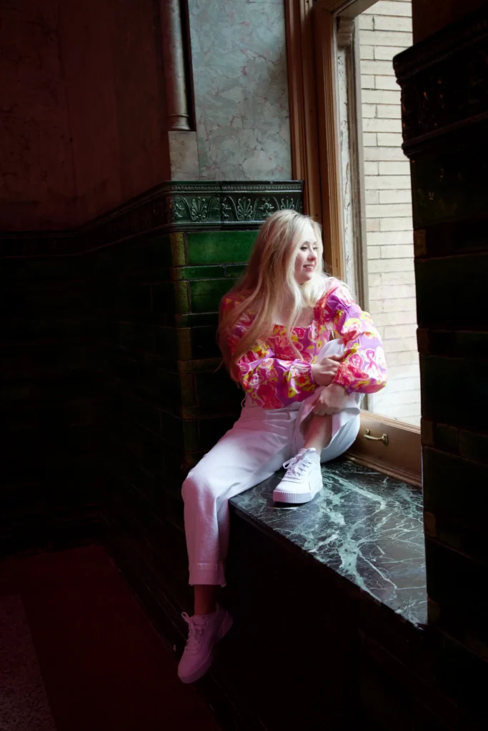 A blonde woman with Down syndrome sitting on a dark marble windowsill in deep shadow, looking out of the window. She is wearing a colorful pink and yellow floral blouse, white pants, and white platform sneakers.