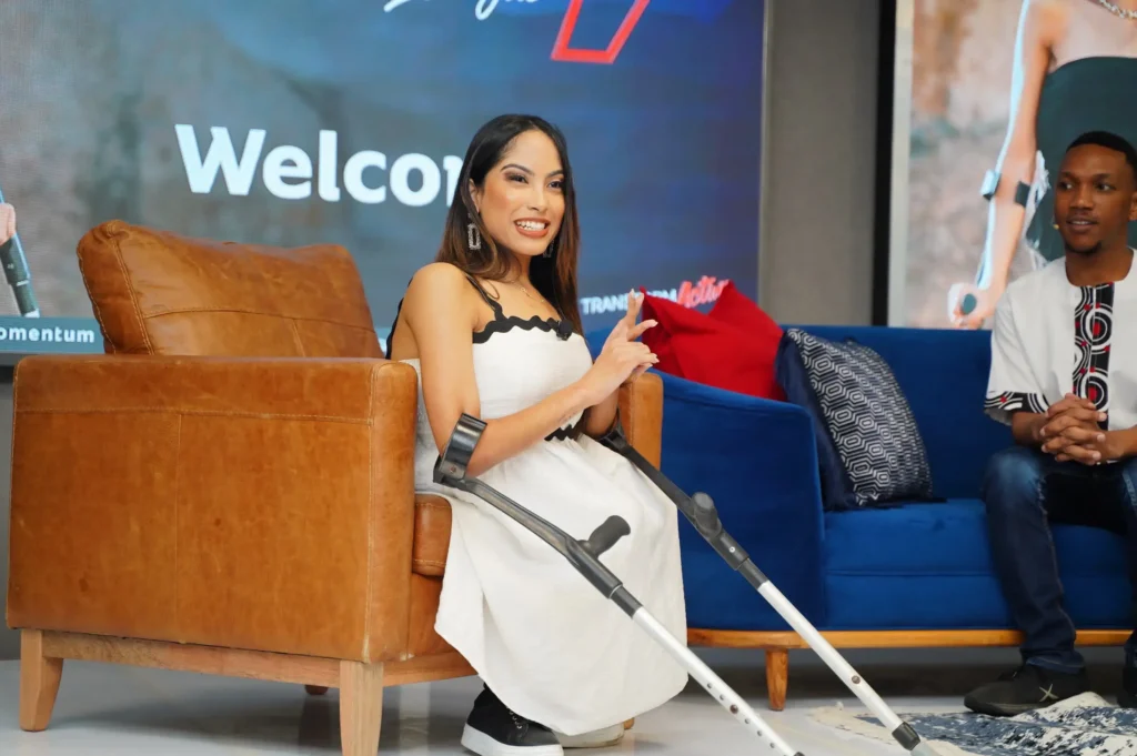 A woman with long dark hair, wearing a white dress with black scalloped trim, is seated on a large brown leather armchair on a stage. She is holding her forearm crutches. A man is seated beside her on a blue velvet sofa. A screen in the background displays "Welcome."