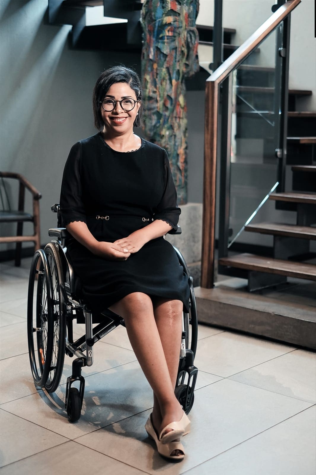 Portrait of a smiling woman wearing glasses and a formal black dress, seated in a wheelchair in a modern indoor space with a wooden staircase and a sculpture in the background. Her hands are clasped in her lap, and she is wearing light-colored, low-heeled shoes.