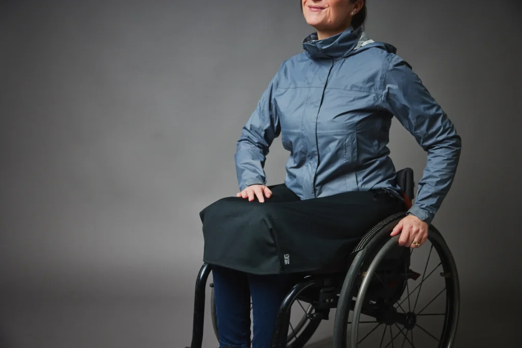 A smiling woman seated in a manual wheelchair against a plain grey background. She is wearing a blue-grey rain jacket and a black, waterproof "Lap Jacket" cover that sits comfortably over her lap and thighs, tucking around her legs for protection against the elements.