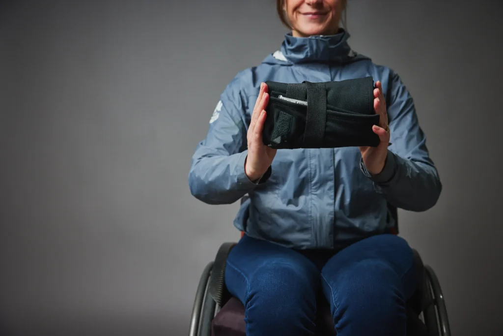 A woman seated in a wheelchair holding up a compact, folded black bundle in her hands. This shows the "Lap Jacket" packed away for storage, secured with a black strap and velcro, demonstrating its portability.
