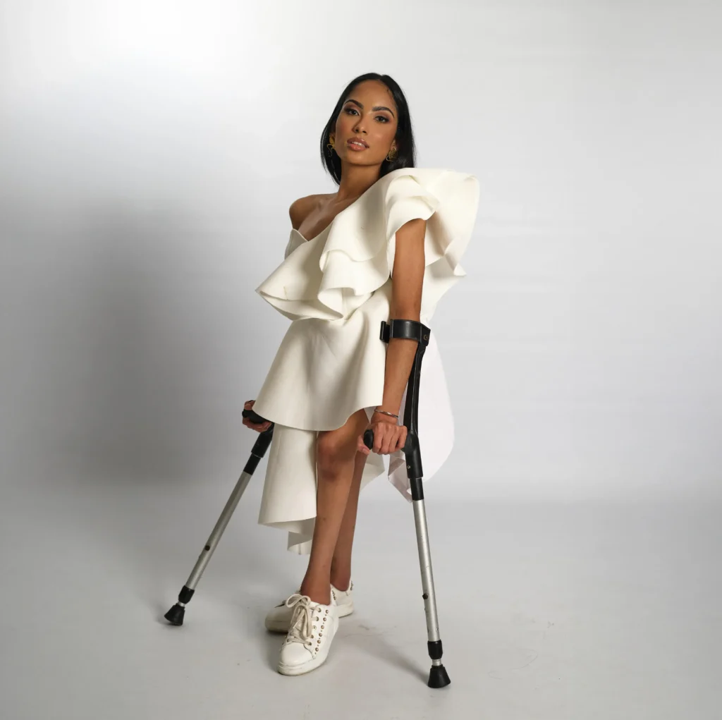 A full-body studio shot of a woman with long dark hair posing against a white background. She is supported by two forearm crutches and wearing a dramatic, asymmetric white dress with a large ruffled shoulder and white sneakers.