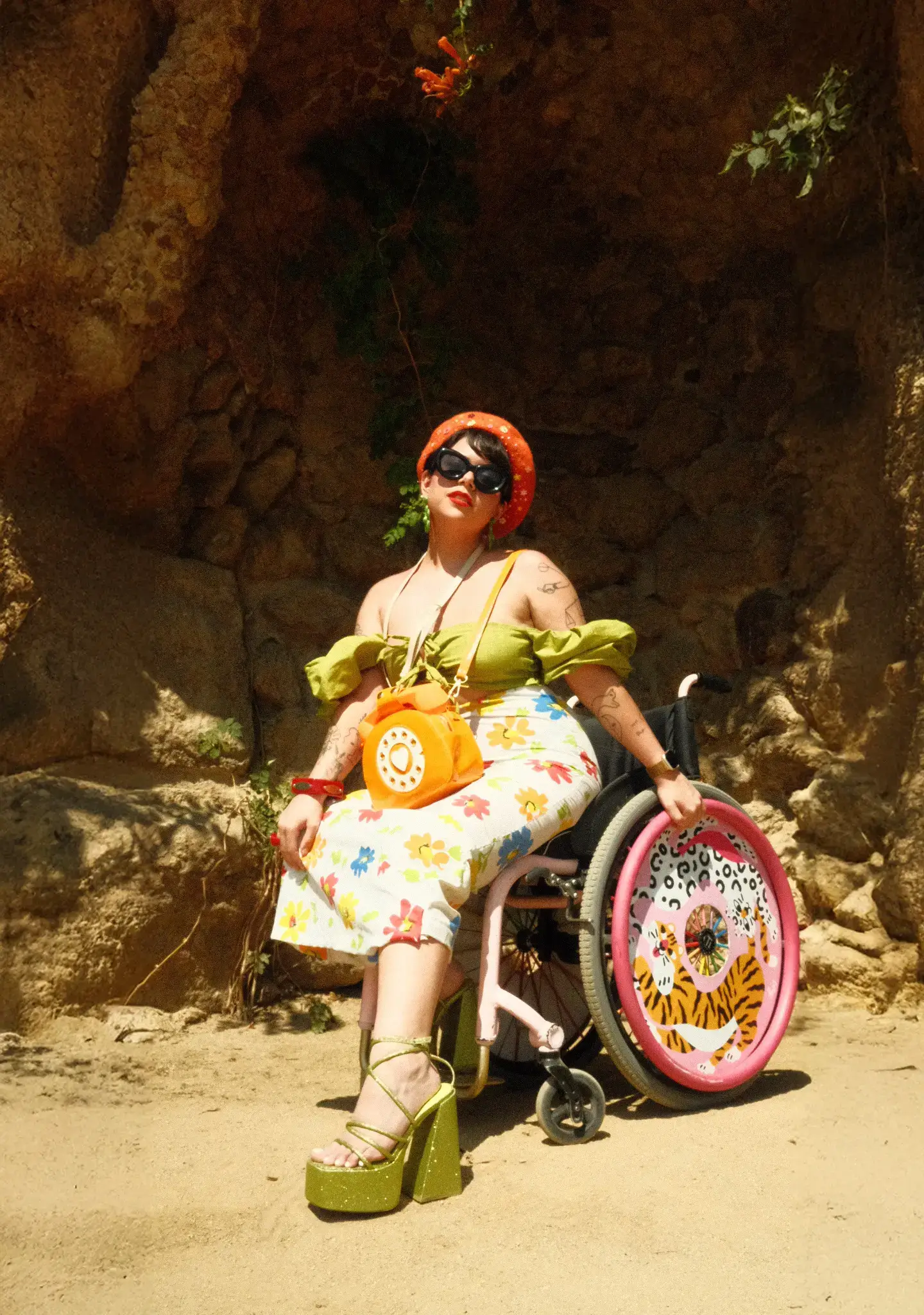 A vibrant photo of a woman seated outdoors in a pink wheelchair against a textured rock wall. She is wearing a green off-the-shoulder crop top, a white floral midi skirt, green platform heels, and an orange beret and sunglasses. A bright orange novelty purse shaped like a rotary phone is slung over her shoulder. The wheelchair's back wheel features a custom pink cover with a stylized tiger design.