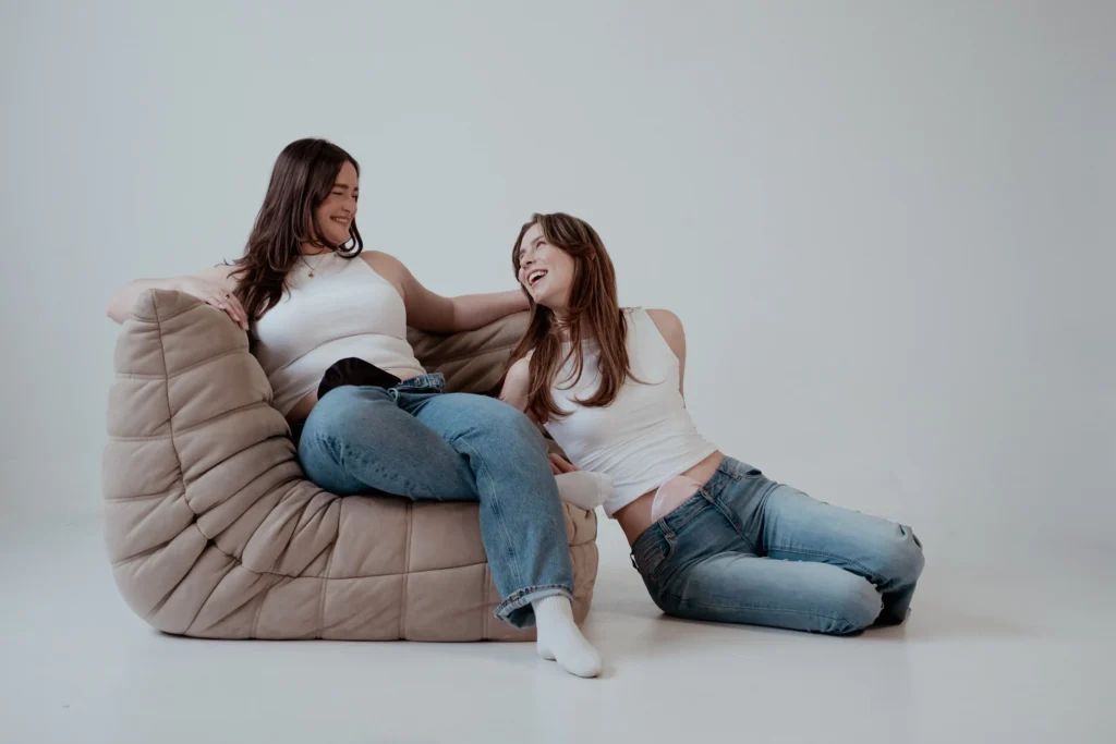 Two young women smiling and laughing together on a beige, overstuffed chair in a bright, white studio. Both are wearing white tank tops and blue jeans; the woman on the left sits on the couch, and the woman on the right leans against it on the floor, showing a small, pale beige ostomy pouch cover visible at her lower waistline.
