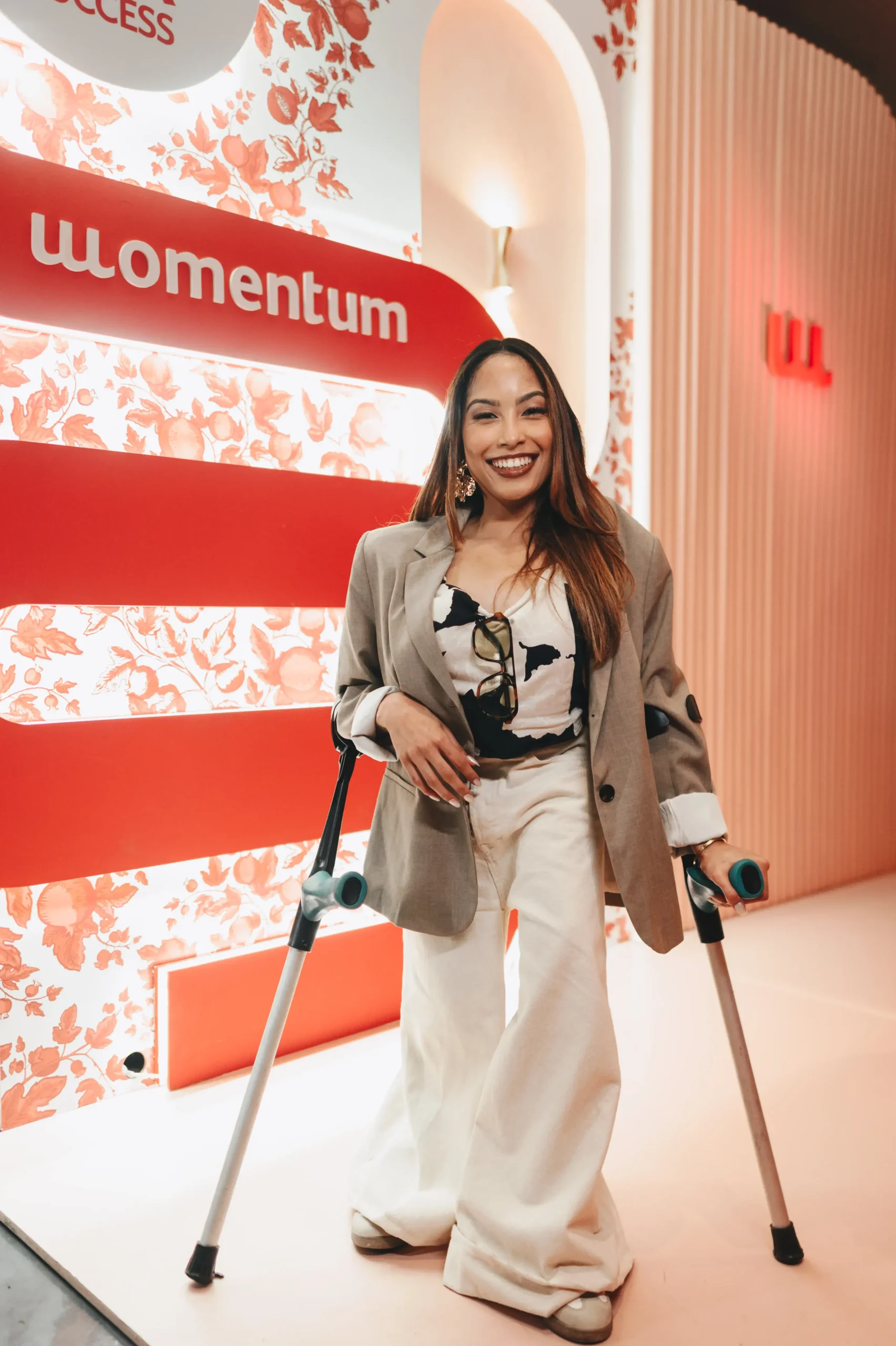 A smiling woman with long dark hair stands indoors, supported by two forearm crutches. She is wearing a modern outfit: a white and black abstract print top, wide-leg off-white trousers, and a relaxed-fit beige blazer. The background is a pink and white archway with a red wall featuring the "Momentum" logo.