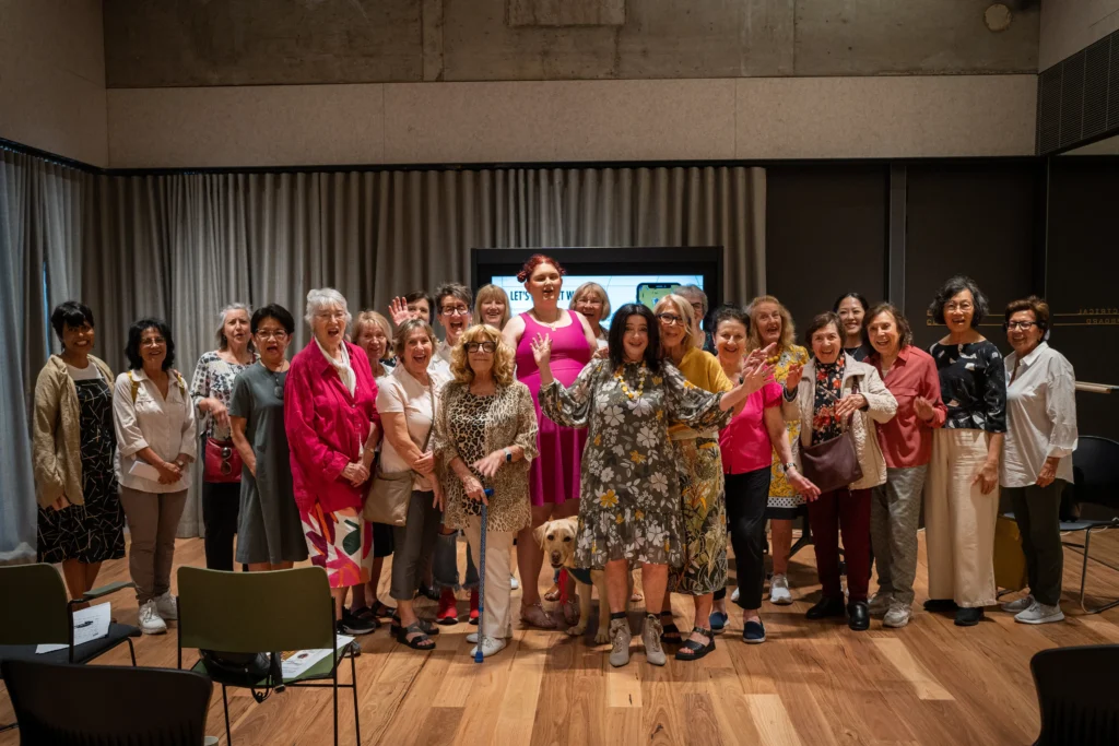 A large group of approximately 20 women of diverse ages are posing together for a group photo in a warmly lit, modern room. They are standing on a polished wooden floor in front of a neutral-colored curtain and a large screen. Many of the women are smiling and waving. One woman in the center is wearing a pink dress, and another nearby is wearing a grey dress with a floral pattern and has a cane and a service dog at her feet.