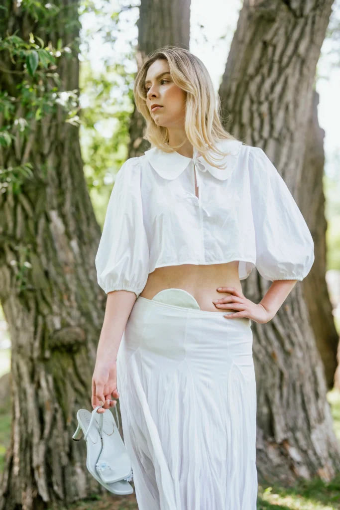 A young blonde woman stands outside between two large tree trunks, holding a pair of white high-heeled shoes in her hand. She is wearing a fashionable white outfit consisting of a cropped, puff-sleeved blouse with a large peter pan collar and a pleated white maxi skirt. A pale beige ostomy pouch cover is visible at her lower abdomen.
