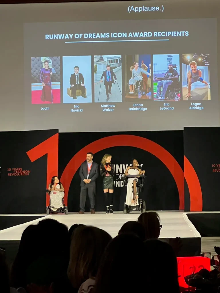 A wide shot of a stage event with an audience in the foreground. A large projection screen displays the text 'Runway of Dreams Icon Award Recipients' along with photos of six honorees. Below the screen, four people—two standing and two using power wheelchairs—are presenting on stage.