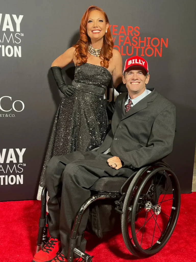 A red carpet photo featuring a woman with red hair wearing a strapless, black sequined gown and pearl necklace standing next to a smiling man in a wheelchair who is wearing a dark suit and a red baseball cap with the name 'BILLY' on it. The backdrop reads 'Runway of Dreams Foundation'.