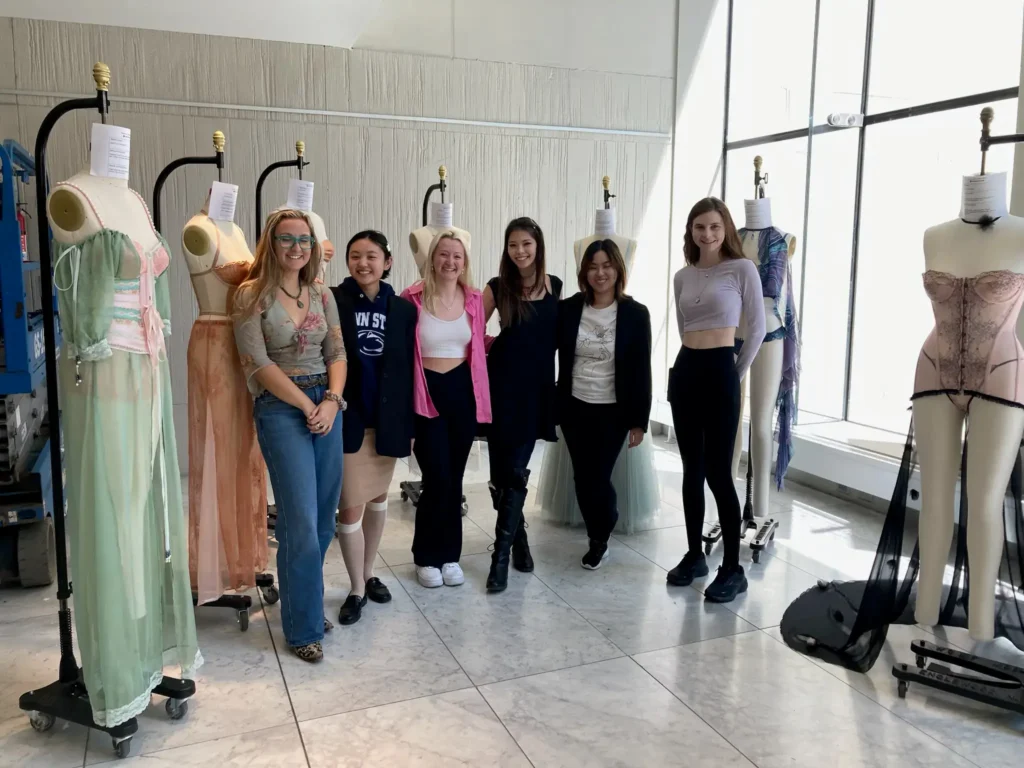 A group of six young women standing and smiling in a bright room with marble floors. They are flanked by dress forms and mannequins displaying custom lingerie and corset fashion designs.