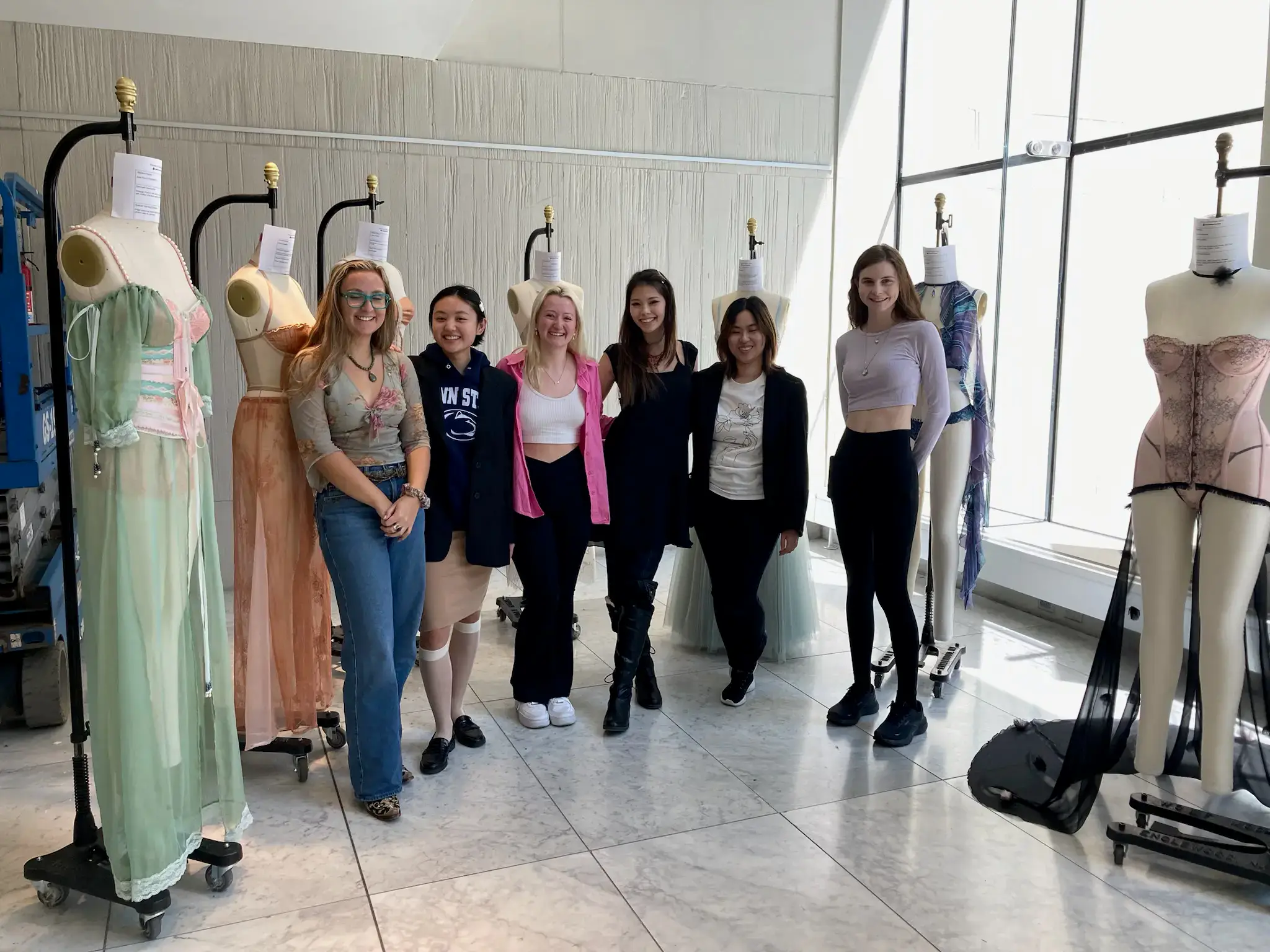 A group of six young women standing and smiling in a bright room with marble floors. They are flanked by dress forms and mannequins displaying custom lingerie and corset fashion designs.