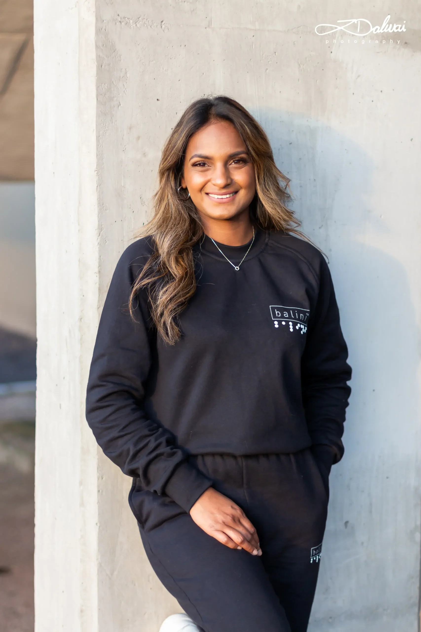 A medium shot of a woman with long, highlighted brown hair smiling while leaning against a concrete wall outdoors. She is wearing a black sweatshirt and matching sweatpants, with a white graphic logo featuring the word "balina" and a series of dots beneath it printed on the left chest area. She also wears a simple silver necklace.