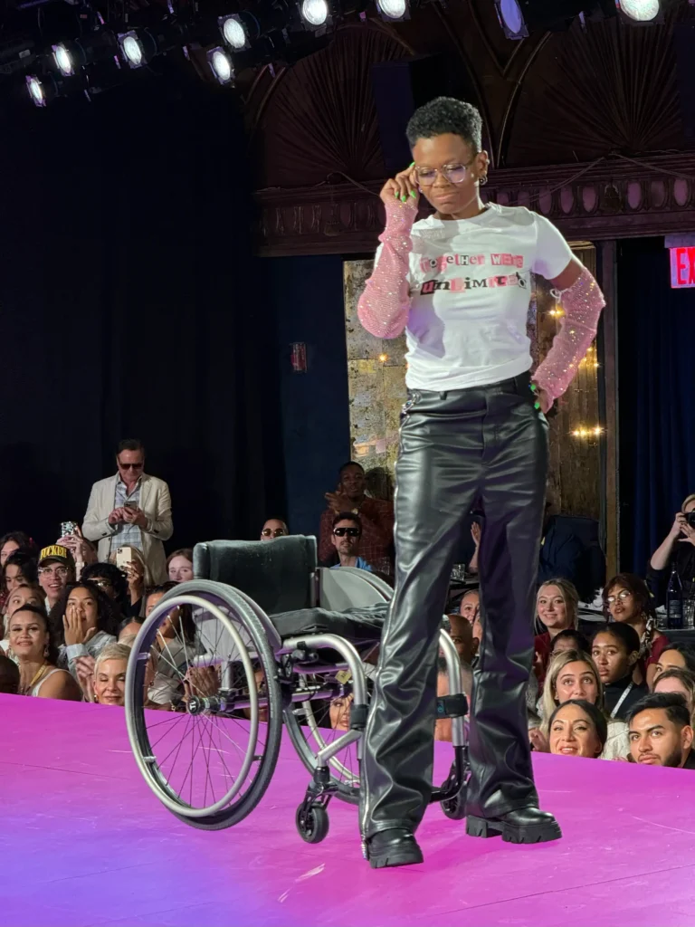 A Black fashion model with short, dark hair and glasses stands proudly next to her silver manual wheelchair on a bright magenta runway. She is wearing a white t-shirt with the text "TOGETHER WE ARE UNLIMIT[ED]," high-waisted black faux leather pants, and sheer, bright pink mesh gloves speckled with glitter. She is looking down and adjusting her glasses. A man in sunglasses is visible clapping in the background audience.