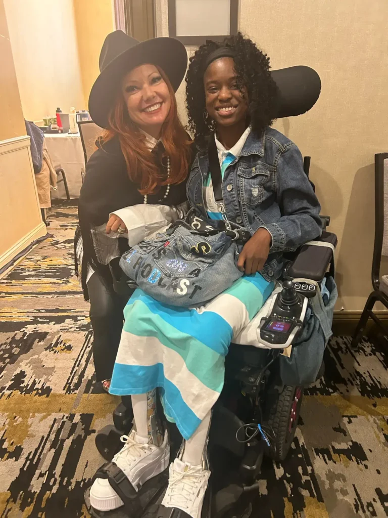 A candid indoor shot of two women smiling. On the left, a woman with red hair wearing a wide-brimmed black hat and fringed poncho leans in next to a young woman seated in a power wheelchair. The woman in the wheelchair wears a denim jacket and a blue-and-white striped dress, holding a denim bag decorated with patches.