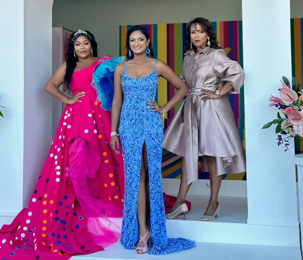 Three women posing under a white archway featuring a "Dove invisible dry" sign with a rainbow-colored circle graphic. The woman on the left wears a flamboyant pink polka-dot gown with blue and turquoise ruffles. The woman in the center wears a floor-length, bright blue, heavily beaded dress with a high slit. The woman on the right wears a sophisticated, knee-length, metallic rosy-gold shirt dress with a wide belt. A display of pink lilies is visible to the right.