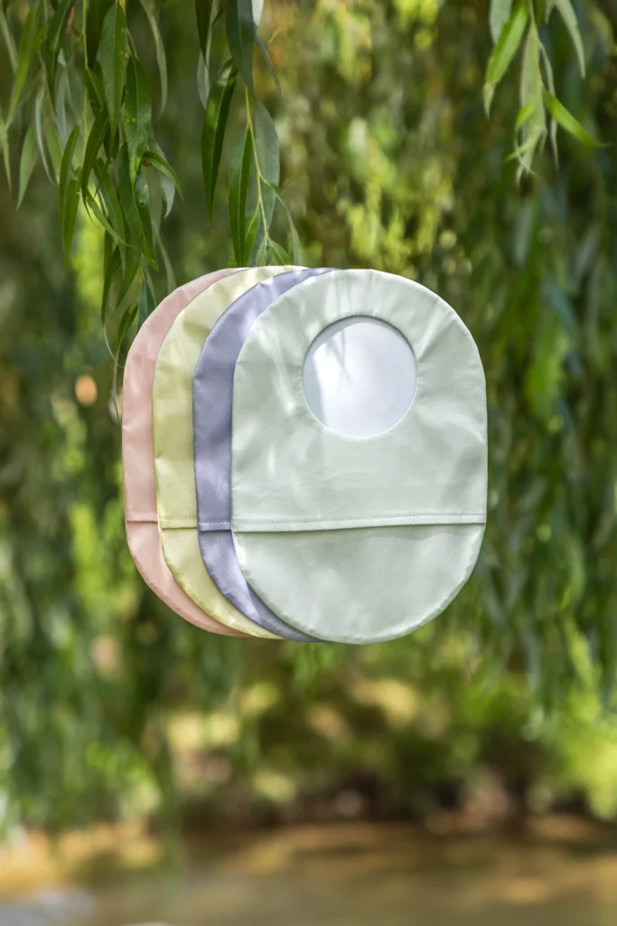 Four colorful, satin-like ostomy pouch covers, stacked and hanging from a thin branch of a weeping willow tree. The colors, from back to front, are dusty pink, pale yellow, light gray-blue, and a muted sage green, which has a smooth white ring visible in the center where the stoma would be. The background is a natural, blurry mix of green foliage and water.