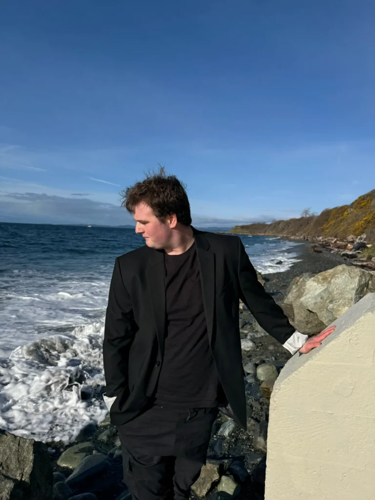 A young man stands on a rocky beach near the water's edge, leaning on a light-colored concrete block. He is wearing a black suit jacket over a black t-shirt and black pants, with the jacket cuffs rolled up. He is looking down towards the water, and the background shows the ocean with white foam and a rocky shoreline backed by a hillside.