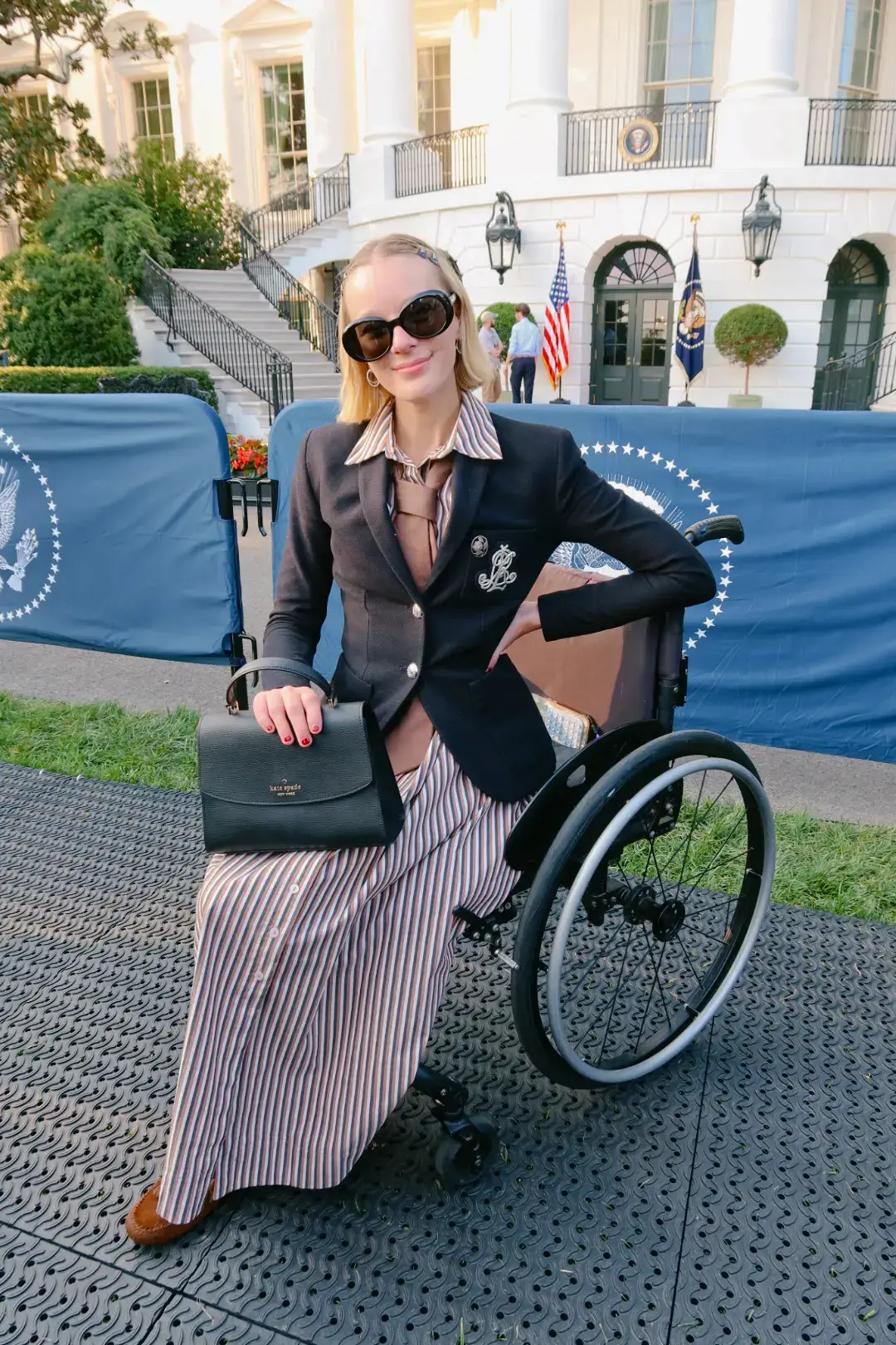 A woman seated in a wheelchair outdoors in front of a white neoclassical building, wearing a tailored black blazer over a striped dress, oversized sunglasses, and holding a black handbag. She poses confidently on a lawn with flags and security barriers visible in the background.