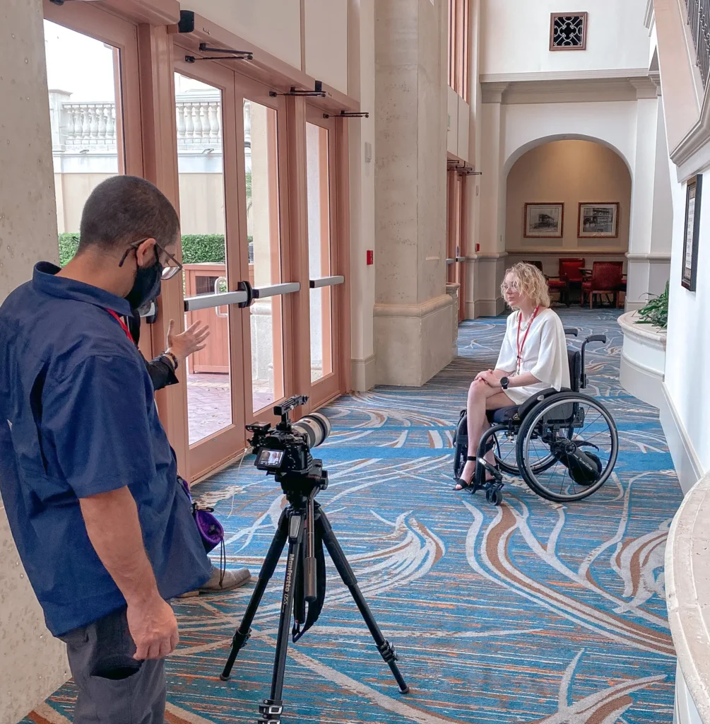 A young woman in a manual wheelchair, wearing a white top and shorts, sitting in a bright, ornate hotel or conference hallway. A man in a blue shirt and black face mask is standing in the foreground on the left, gesturing while looking toward a camera set up on a tripod, seemingly preparing for a video shoot. Large wooden doors are visible behind him, and the floor is covered in a blue patterned carpet.