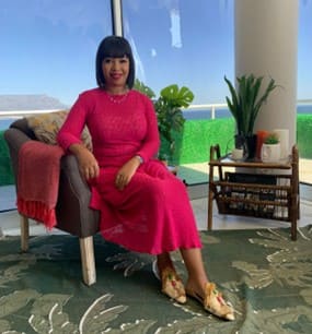 A stylish woman with a dark bob haircut is seated in an armchair by a large window overlooking a coastal city skyline, likely Cape Town's Table Mountain. She is wearing a fitted, bright pink long-sleeved dress and ornate, patterned slip-on shoes.