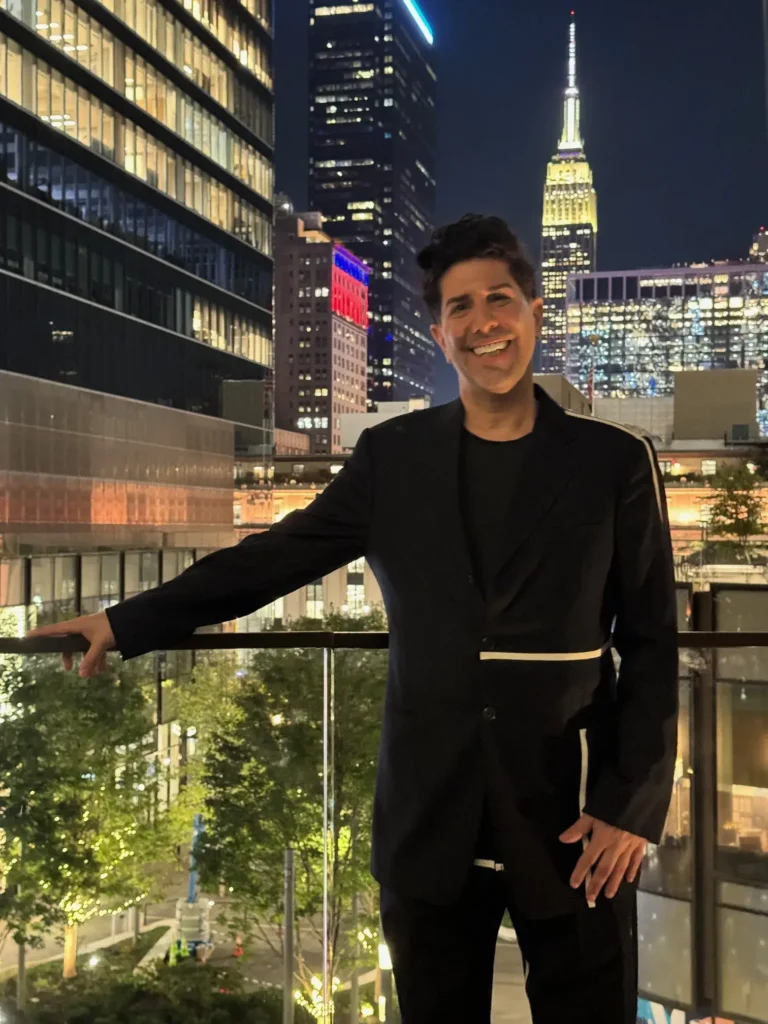 A photograph of Ramy Gafni, a dark-haired man with a mustache and soul patch, smiling outdoors at night. He is wearing a dark suit jacket with a white horizontal stripe and stands on a balcony with a glass railing. In the background, there is a city skyline with tall buildings lit up, including the Empire State Building with its iconic spire visible.
