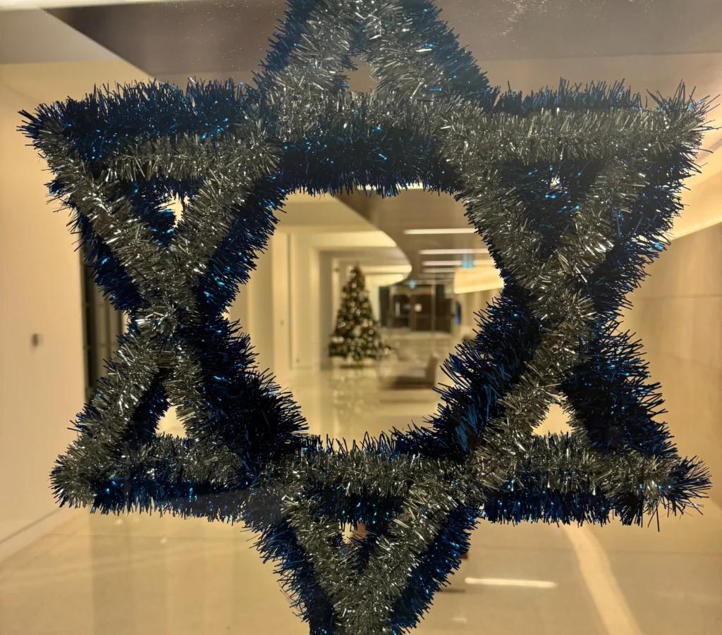 A close-up of a Star of David (Magen David) decoration made of shimmering blue and silver tinsel, attached to a glass window or door. The center of the star is transparent, framing a blurred view of a modern, brightly lit hallway in the background where a decorated holiday tree is visible in the distance.