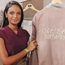 A close-up portrait of a woman smiling, standing next to a jacket hanging on a rack. The woman, with dark hair and a maroon top, has her hand resting on the sleeve of a pinkish-tan jacket. The back of the jacket features a horizontal design composed of raised white dots, which appear to be Braille symbols, visible across the back shoulder area.