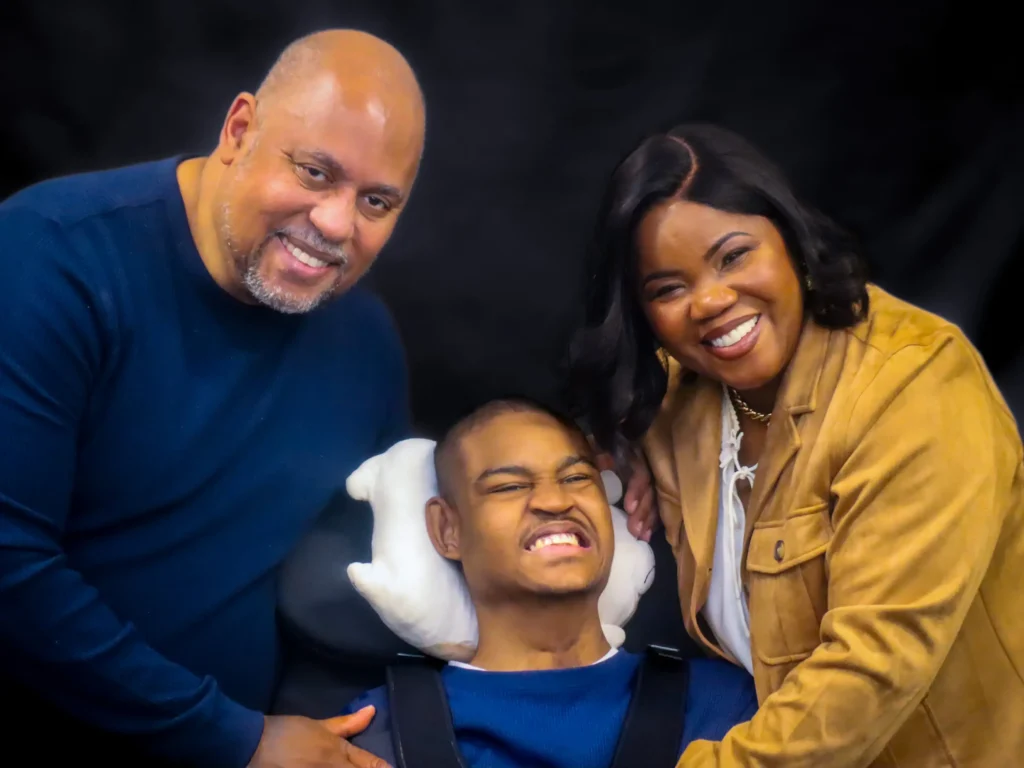 A heartwarming studio portrait of a Black family against a dark background. A man in a blue sweater and a woman in a tan jacket lean in smiling toward a young man seated in the center, who is smiling broadly and using a white supportive headrest.