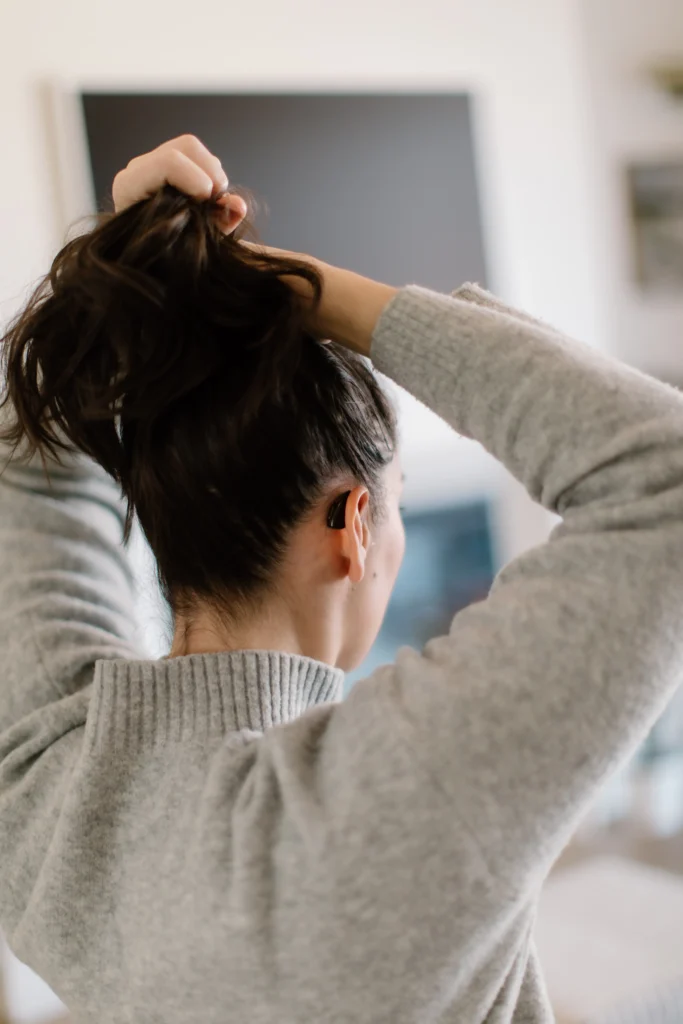 r view of a woman with her dark hair gathered up in her hands as if she is about to tie it. A black behind-the-ear hearing aid is clearly visible on her right ear. She is wearing a grey ribbed sweater.