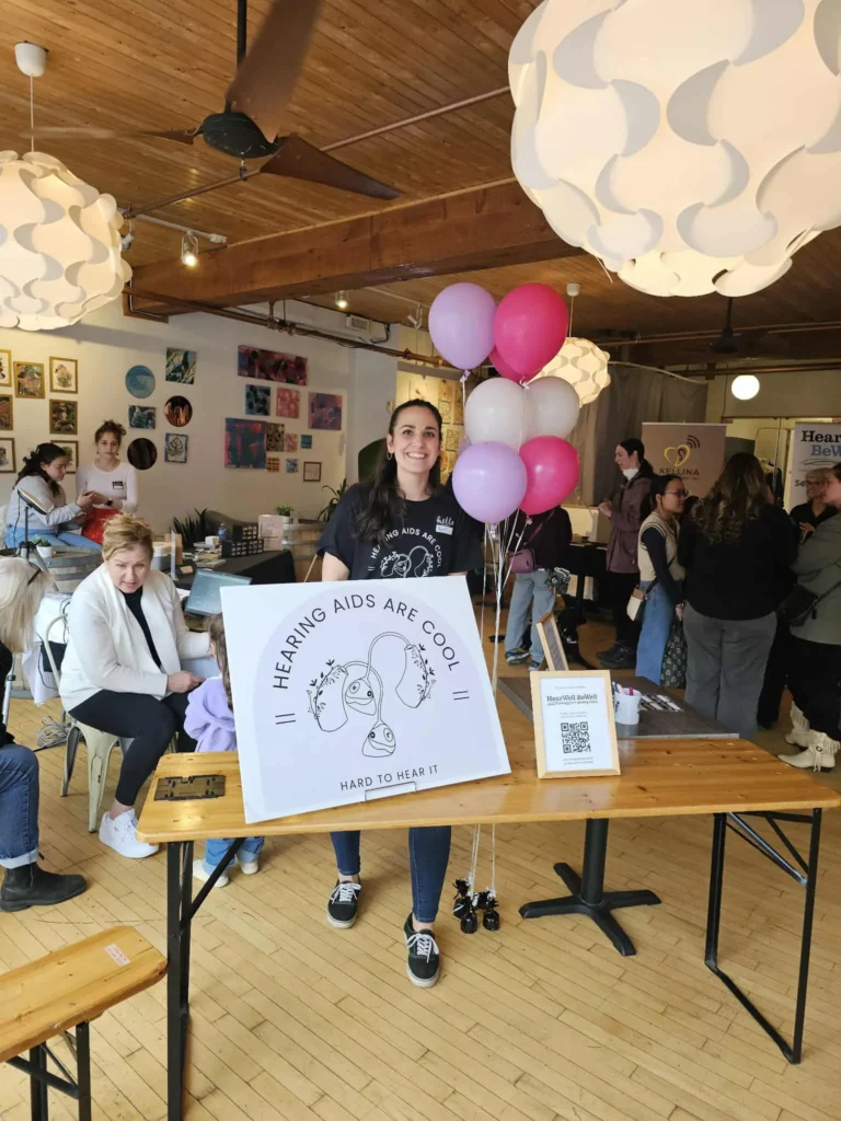A woman stands smiling behind a wooden table at an event. On the table is a large sign that reads "HEARING AIDS ARE COOL" with an illustration of ears. She is surrounded by pink and white balloons, and the background shows a busy community space with art on the walls and other people.