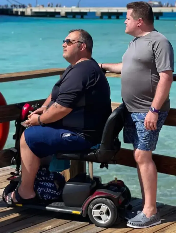 Two men, Matty and Scott, enjoying a sunny day on a wooden pier by bright turquoise water. Matty is seated on a red mobility scooter wearing a black t-shirt and blue shorts, looking out at the ocean. Scott stands beside him in a gray t-shirt and patterned blue shorts, resting a hand on the scooter.