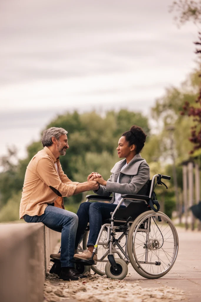 A middle-aged man with salt-and-pepper hair sits on a low stone wall outdoors, tenderly holding both hands of a Black woman sitting in a wheelchair. They are looking into each other's eyes and smiling warmly against a soft-focus background of green trees and a pale sky.