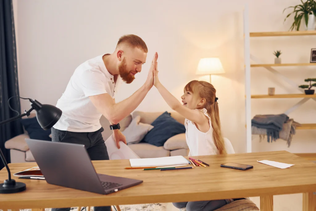A father with a red beard and his young daughter share an enthusiastic high-five over a wooden desk. A laptop, colored pencils, and papers are spread across the desk. The scene is set in a warm, brightly lit living room with a couch and shelving unit in the background.