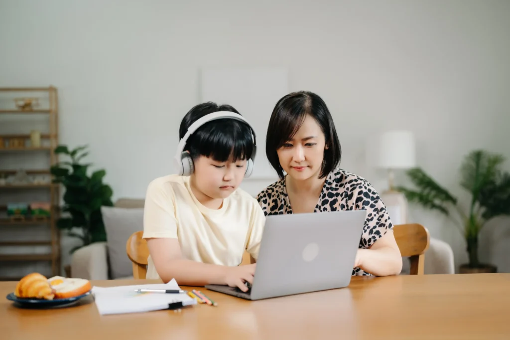 An Asian woman in a leopard-print blouse sits at a wooden table next to a young boy. The boy is wearing white over-ear headphones and focusing on a silver laptop. A plate with a croissant and eggs, along with some colored pencils, sits on the table in a bright, homey setting.