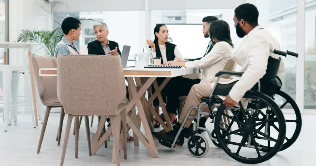 A diverse group of colleagues engaged in an active discussion around a large white conference table in a modern, bright office. The group includes people of various ethnicities and ages, including a man in the foreground using a black wheelchair.