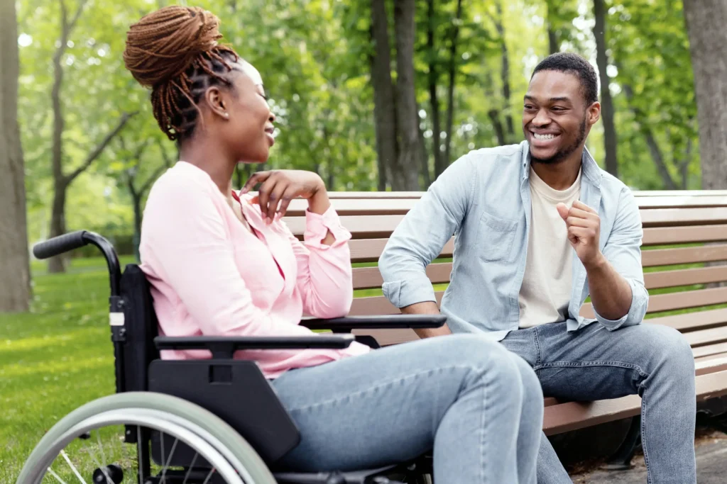 A young Black woman with her hair styled in a high braided bun sits in a wheelchair in a park, laughing and talking with a young Black man sitting next to her on a wooden bench. The man is smiling broadly and gesturing with his hand as they enjoy a bright, sunny day.