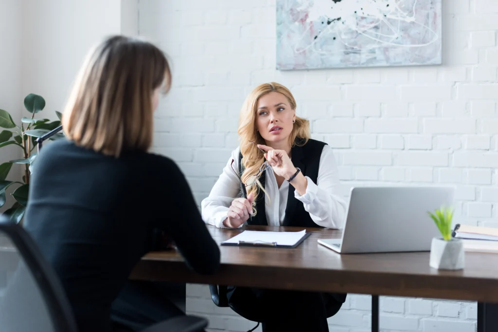 A blonde woman in a white blouse and black vest sits at a wooden desk in a bright office, holding her glasses and speaking with a focused expression. She is facing a colleague, seen from the back, who is wearing a black sweater. A laptop and documents are on the desk between them.