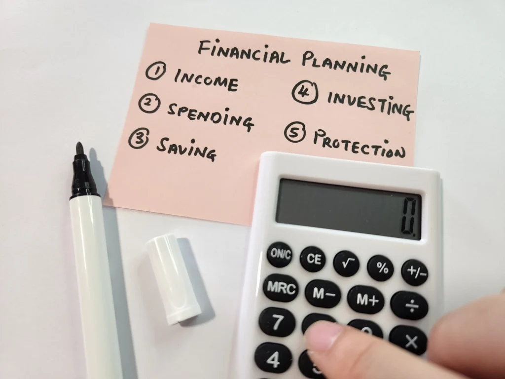 A close-up of a hand using a white calculator next to a pink sticky note. The note lists five numbered points of financial planning: Income, Spending, Saving, Investing, and Protection. A black marker lies nearby.