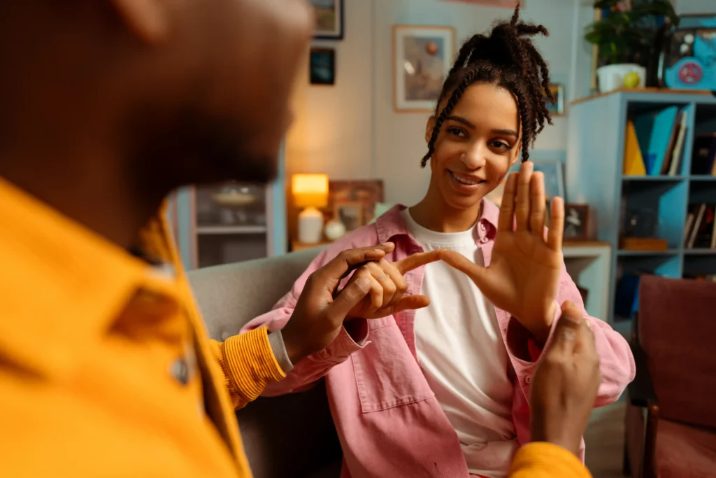 A warm, indoor scene showing two people interacting. A woman with her hair in locs smiles while using her hands to communicate, touching the hand of a man who is partially visible in the foreground. The lighting is cozy and soft, suggesting a personal and inclusive conversation.