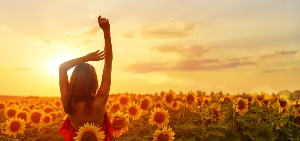 A wide landscape shot of a woman seen from behind in a field of sunflowers. She is wearing a red dress and has her arms raised toward the golden sunset sky, symbolizing freedom and connection with nature.